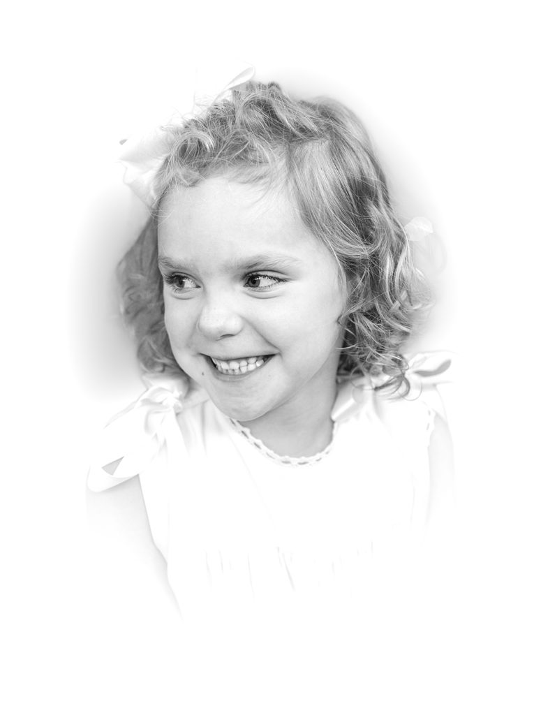 Black and White Heirloom Portrait little girl wearing a white dress with ecru ribbon smiling over her shoulder