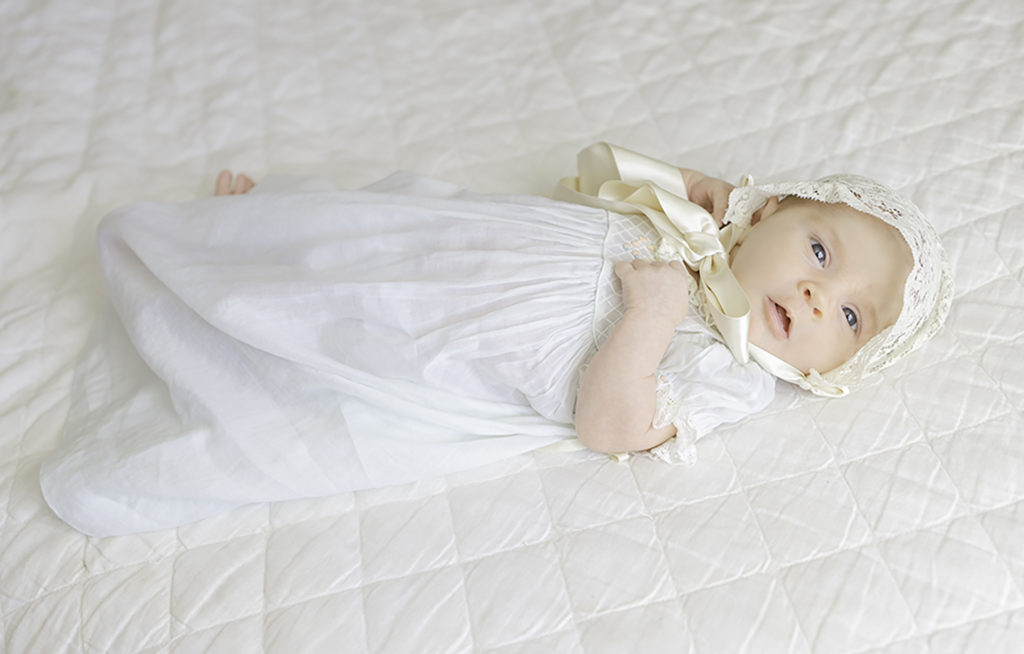 Newborn Baby in daygown with lace bonnet lying on the bed by Birmingham Newborn Photographer