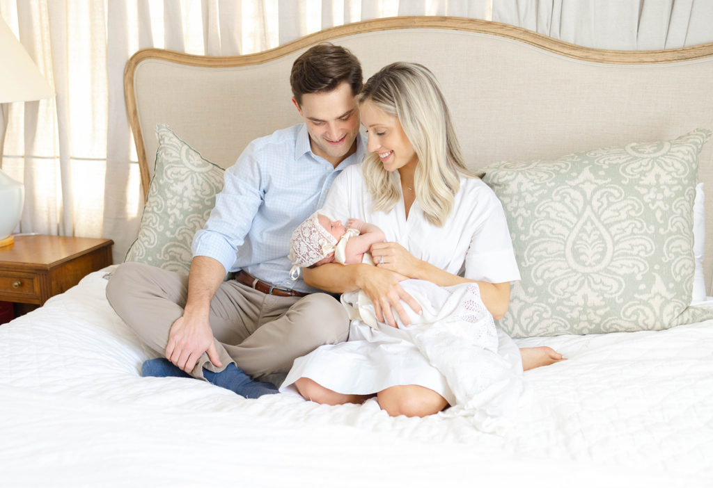 Parents sitting on bed holding and smiling at their newborn baby wearing a bonnet and a swaddle by Birmingham Newborn Photographer Whitney Carr Photography 