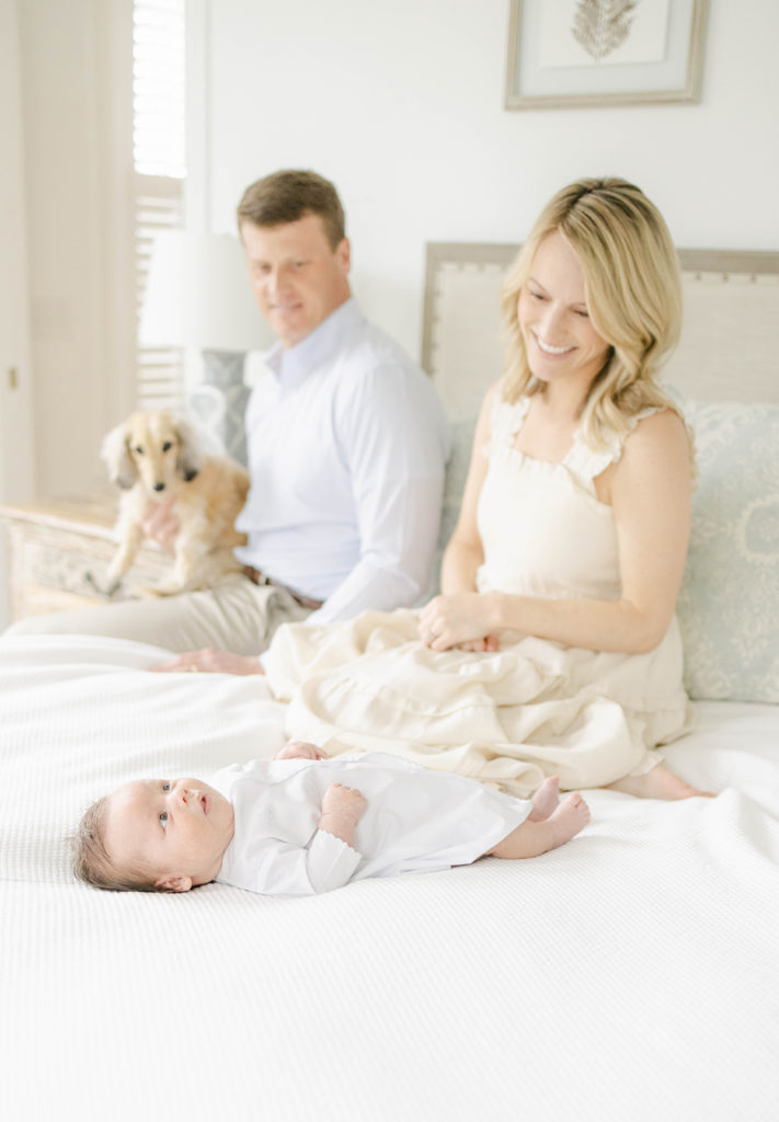 Mom, Dad, and dog sitting on the bed looking down at newborn baby boy lying on bed by Mountain Brook newborn photographer 