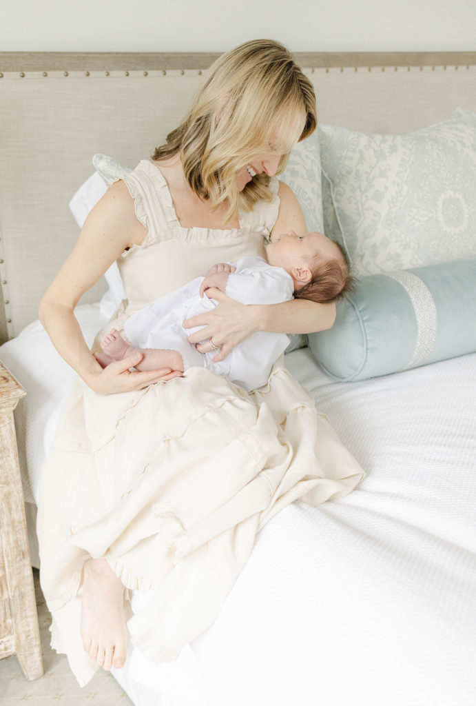Mom wearing a neutral dress and sitting on the edge of the bed holding and looking down at her newborn baby boy wearing a daygown by Mountain Brook newborn photographer 