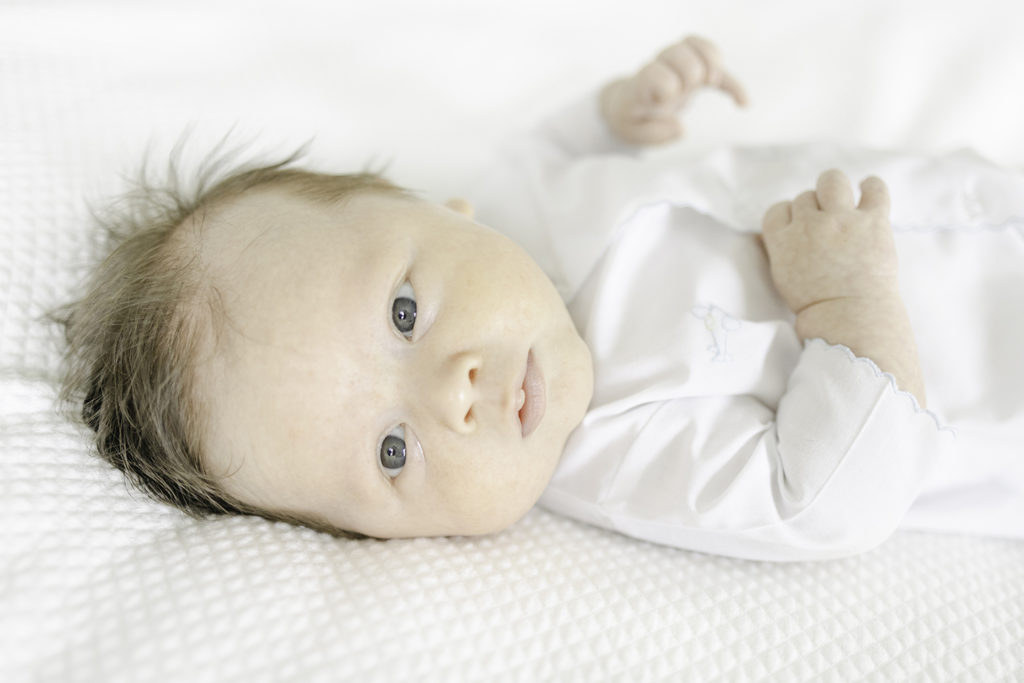Newborn baby boy in Auraluz daygown lying on white bedding and looking at the camera by Mountain Brook newborn photographer 